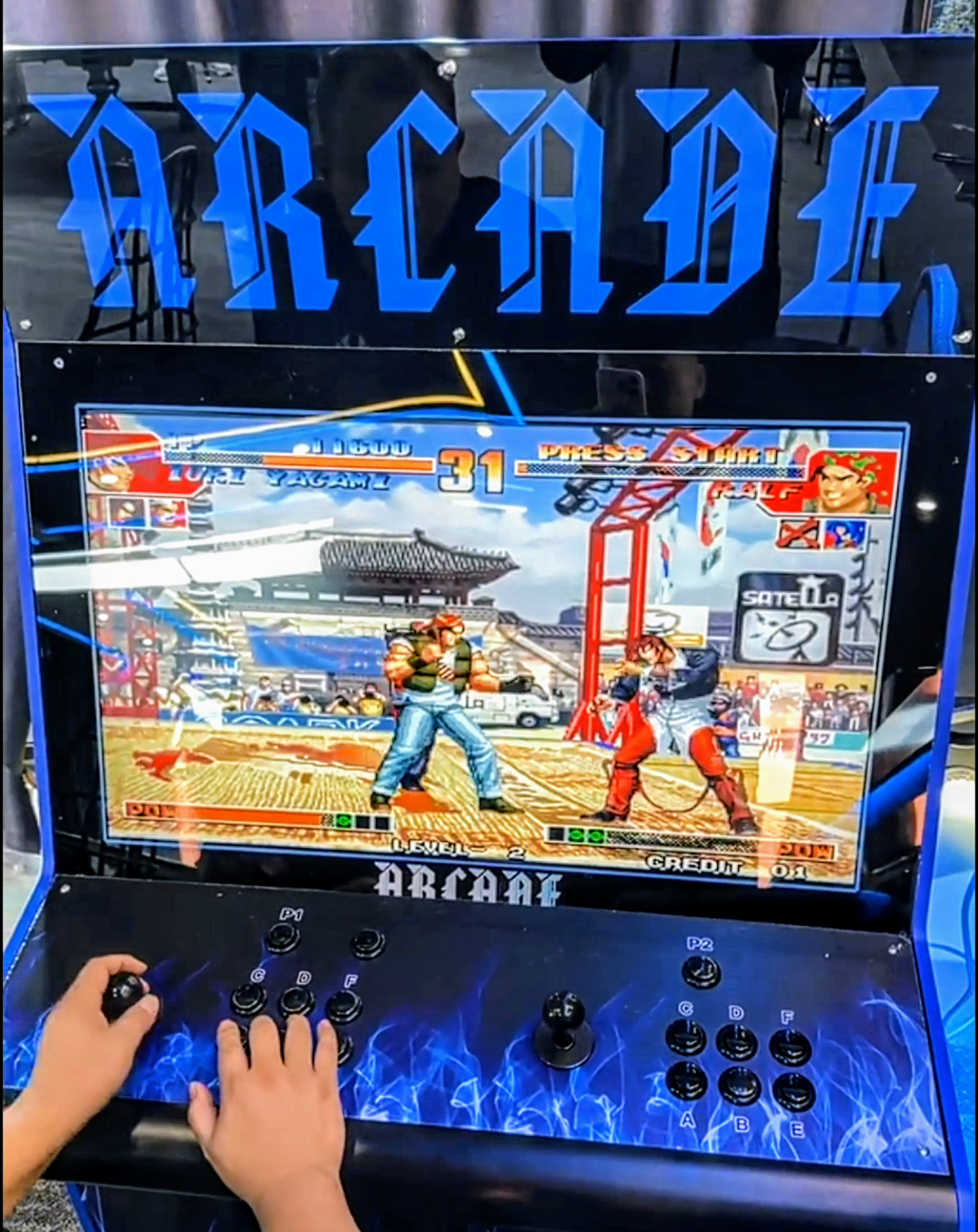 Person playing the game street fighter on a 2 player arcade cabinet with big blue lettering on the top that says ARCADE. The arcade cabinet is black with vibrant blue flames.